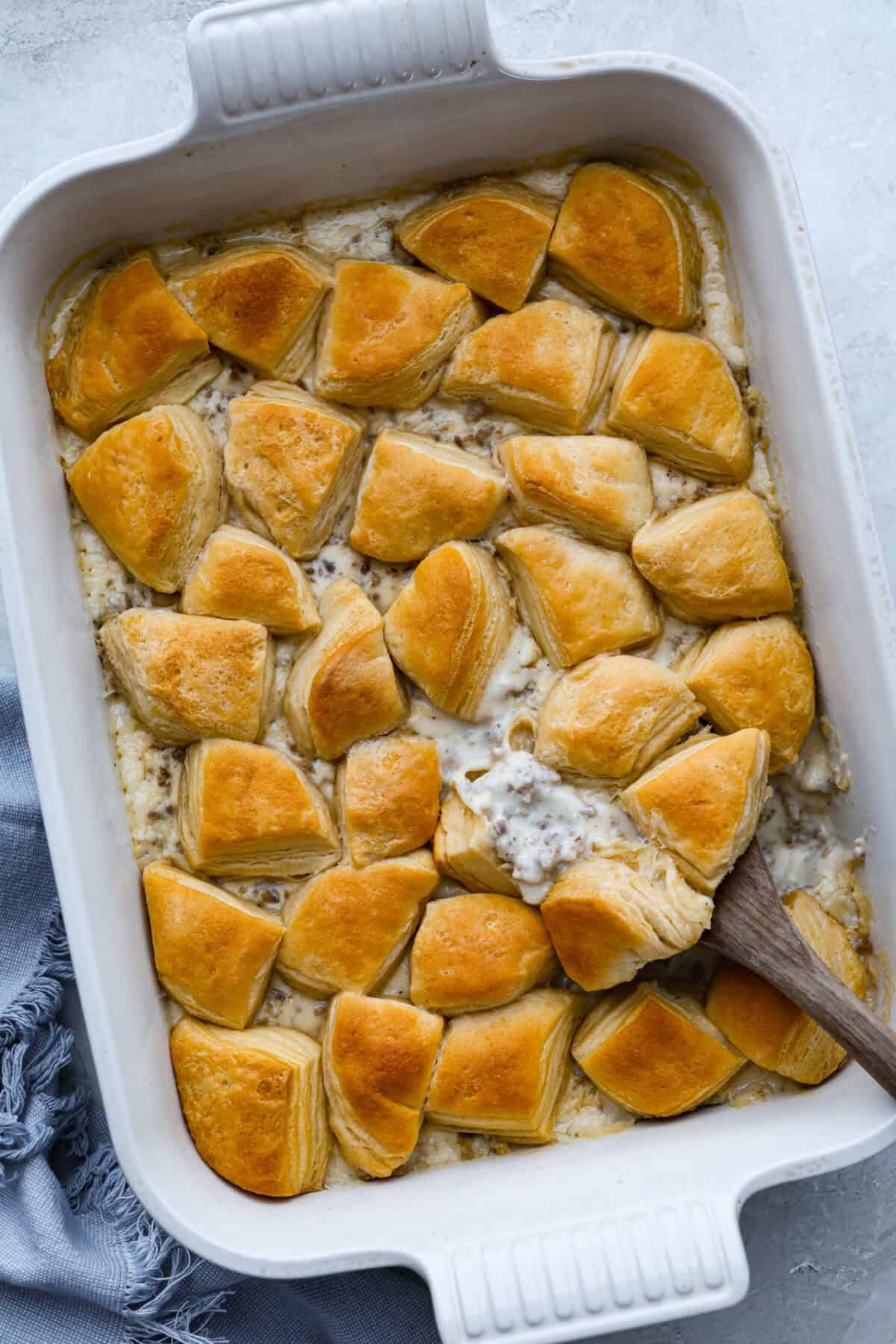 Casserole dish with biscuits and gravy casserole baked to perfection and a wooden spoon taking a scoop out.