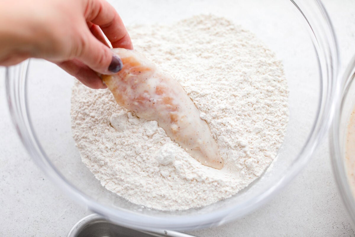 Someone dipping a marinated chicken tender into the bowl of breading ingredients. 