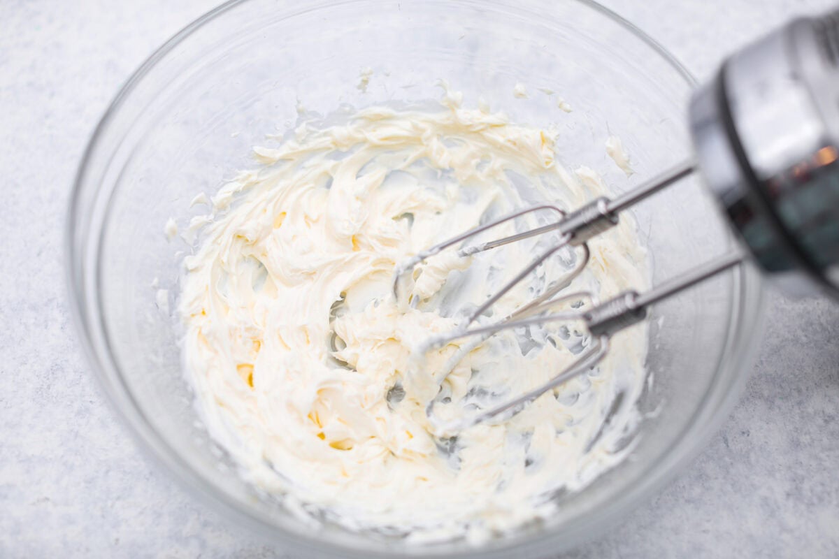 Cream cheese being beaten with a hand mixer in a mixing bowl. 