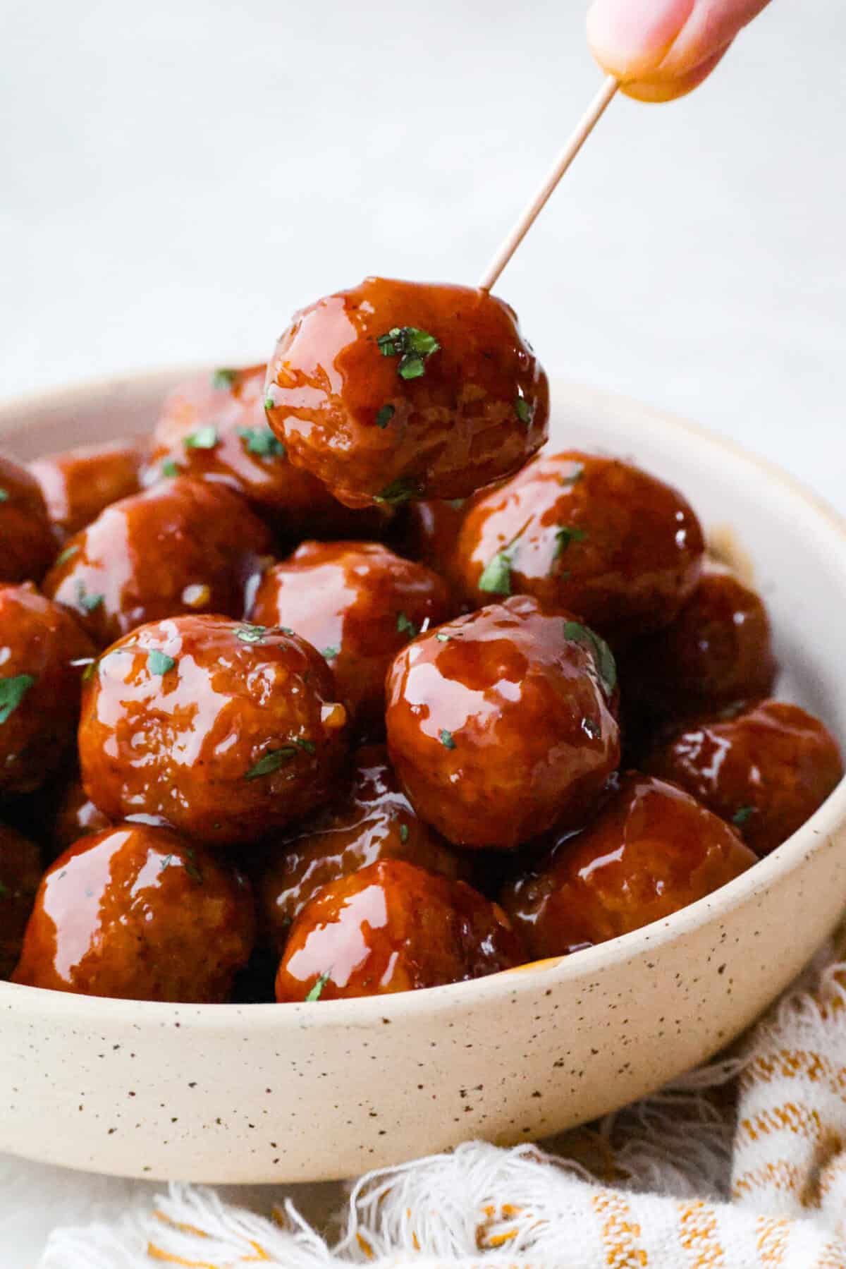Someone using a toothpick to pick up a slow cooker bourbon meatball out of a bowl. 