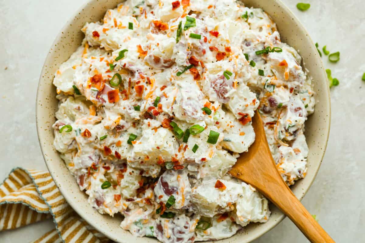 You guys have been asking, so I made the change. This Crack Potato Salad now has mayonnaise in the dressing! It seriously takes the flavors up a notch! I love the combination with the sour cream. I can't wait for you to try this new version! Overhead shot of crack potato salad all mixed up in a serving bowl with a wooden spoon.