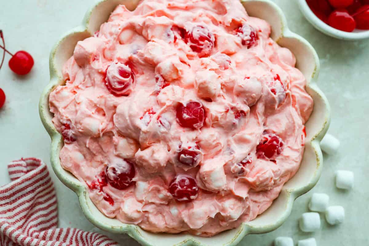 Fluff salads never go out of style. They're light, creamy, and just plain fun! This cherry cheesecake fluff is customizable and so easy to throw together! It's packed with sweet-tangy cheesecake flavor and juicy cherries in every bite. Bowl of cherry cheesecake fluff salad with cherries and marshmallows on the side.